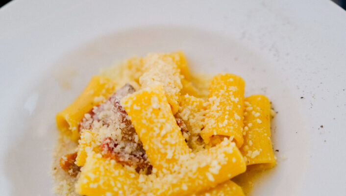 Carbonara: A white plate filled with rigatoni pasta, topped with grated cheese and bits of bacon, sits on a dark table. Another plate of pasta is blurred in the background.