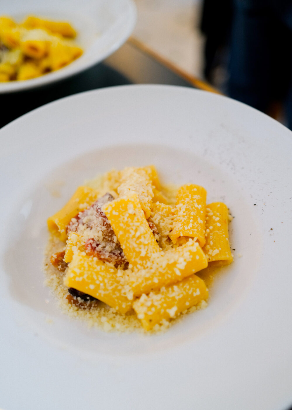 Poiché non è stato fornito alcun titolo per l'immagine nel prompt, il testo alternativo appropriato senza titolo sarebbe: Un piatto bianco con rigatoni conditi con formaggio grattugiato e pezzetti di pancetta o pancetta. Un altro piatto è parzialmente visibile sullo sfondo. Se fosse stato inserito un titolo per l'immagine, ad esempio "Rigatoni alla Carbonara Classica", il testo alternativo sarebbe stato formattato come: Rigatoni alla Carbonara Classica - Un piatto bianco con rigatoni conditi con formaggio grattugiato e pezzetti di pancetta o pancetta. Un altro piatto è parzialmente visibile sullo sfondo.
