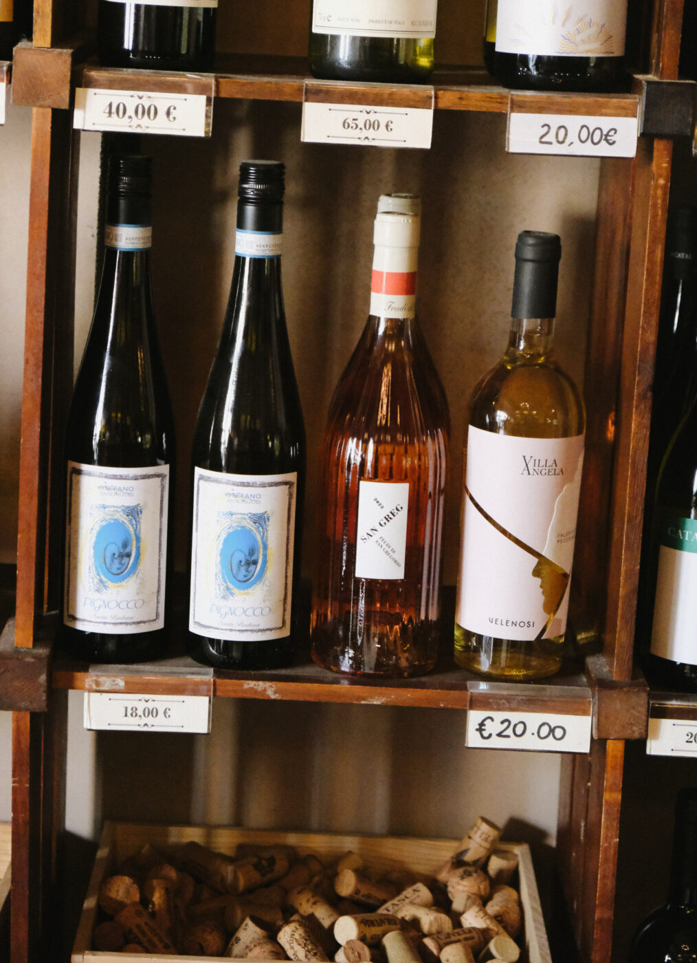 Bottiglie di vino su scaffali di legno - Bottiglie di vino esposte su scaffali di legno, ognuna con etichette diverse e prezzi che vanno dai 18 ai 65 euro. I tappi di sughero sono raccolti in uno scomparto sotto gli scaffali.