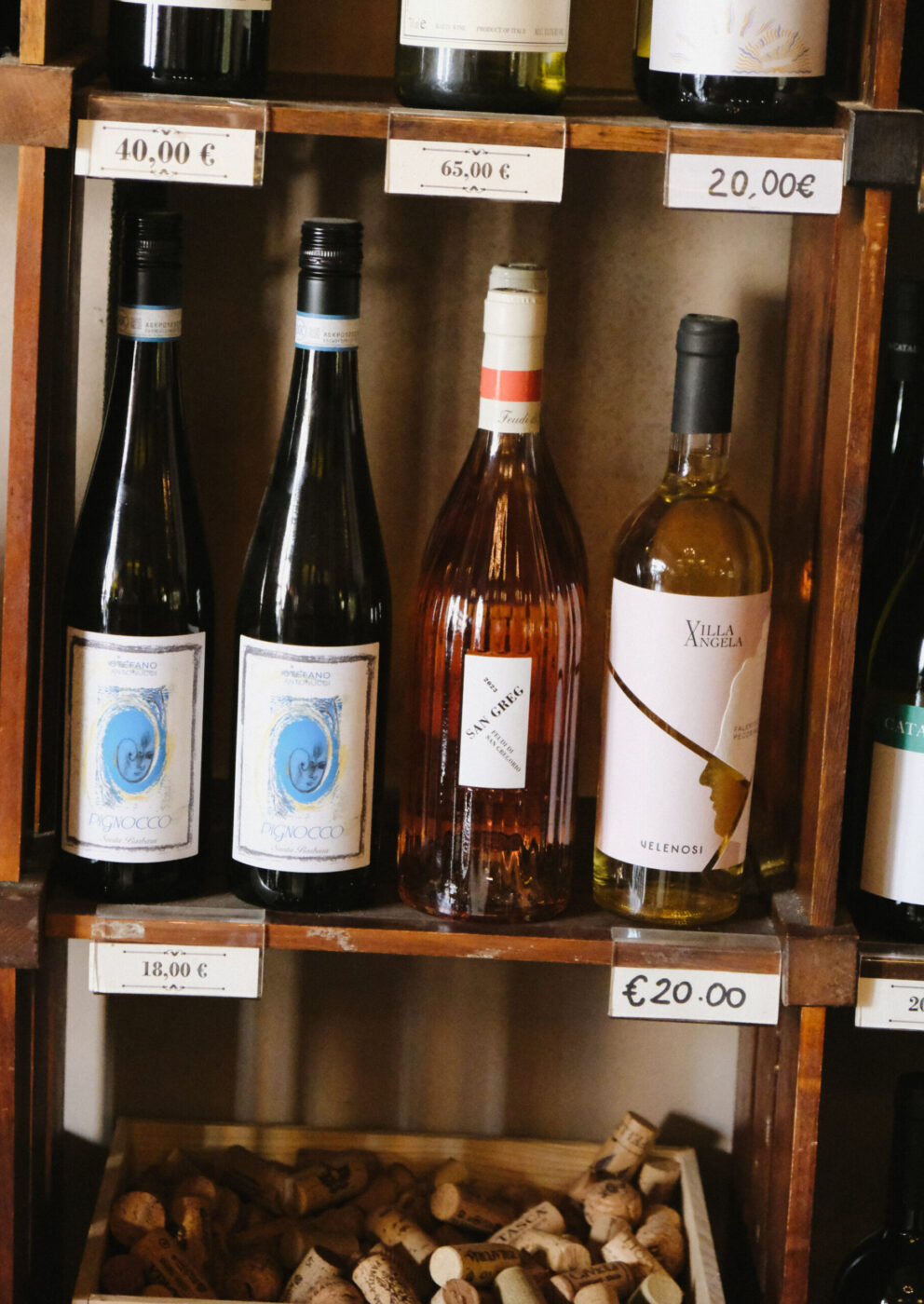A wooden shelf displays bottles of wine with price tags, including sparkling wine, rosé, and white wine; a collection of corks is visible in a compartment below the bottles.