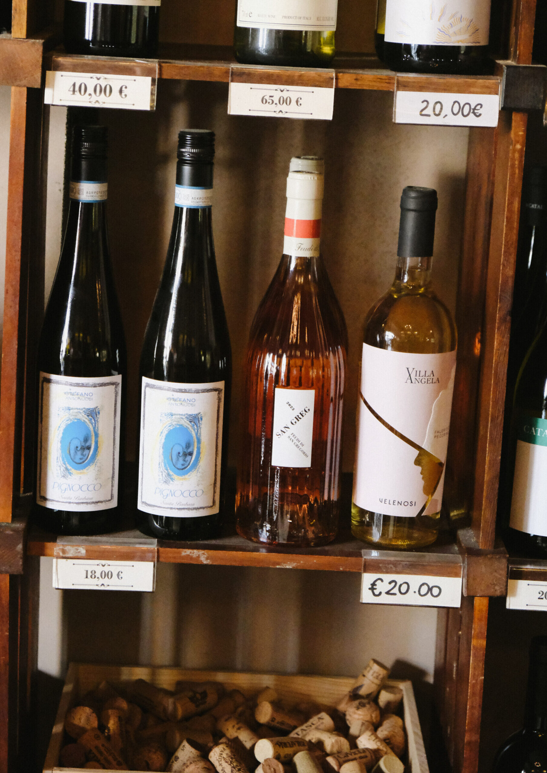 A wooden shelf displays bottles of wine with price tags, including sparkling wine, rosé, and white wine; a collection of corks is visible in a compartment below the bottles.