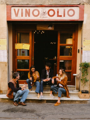 VINO OLIO - Four people sit on the steps outside a shop named VINO OLIO, drinking bottled beverages and talking. The storefront has a wooden door, large windows, and yellow posters on either side of the entrance.