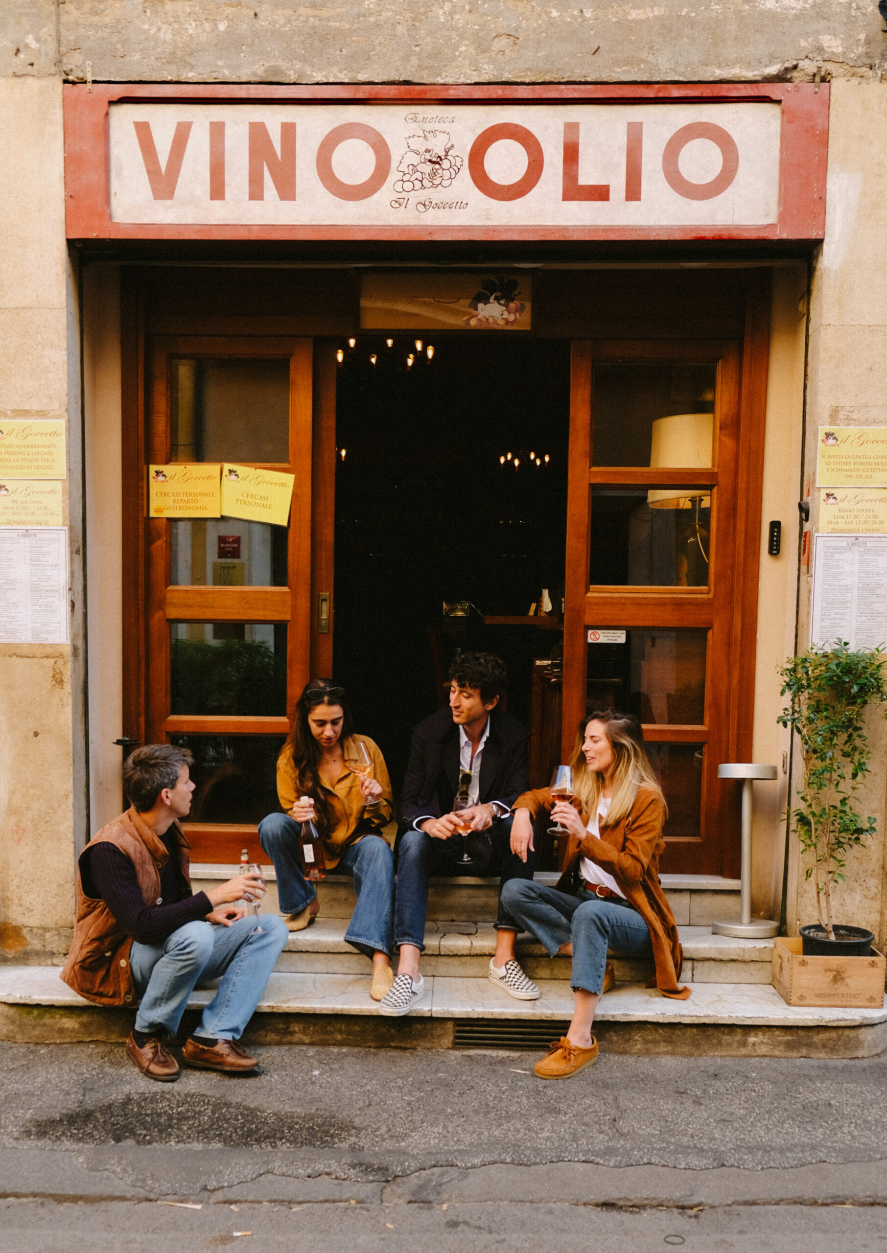 VINO OLIO - Four people sit on the steps outside a shop named VINO OLIO, drinking bottled beverages and talking. The storefront has a wooden door, large windows, and yellow posters on either side of the entrance.