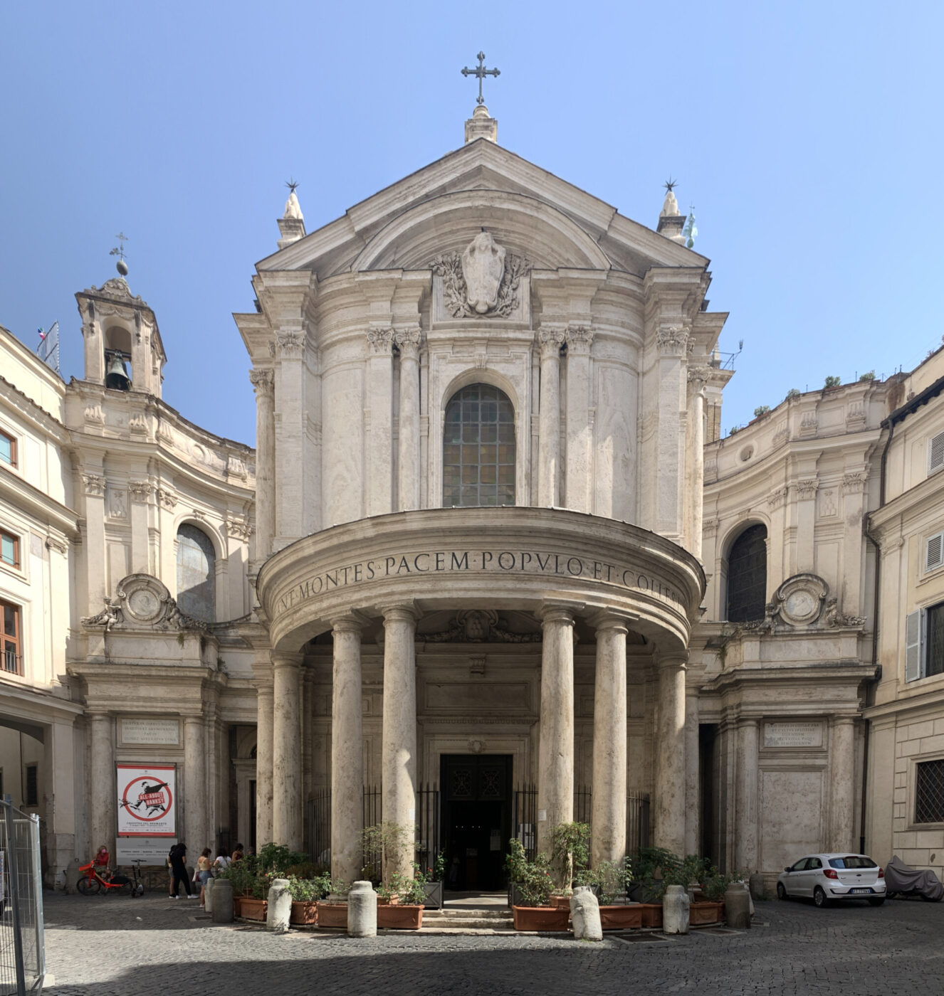 Facciata di una chiesa in stile barocco - Chiesa in stile barocco con alte colonne, un portico arrotondato, un ingresso ad arco e un'iscrizione latina sopra il portale. Una croce sormonta la facciata. Automobili e piante si trovano di fronte, con edifici adiacenti su entrambi i lati.
