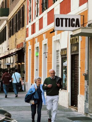 OTTICA - Una coppia di anziani cammina lungo un marciapiede cittadino, passando davanti a edifici colorati, tra cui uno con un cartello con la scritta "OTTICA". L'uomo legge un giornale mentre la donna porta una borsa. Altri pedoni sono visibili sullo sfondo.