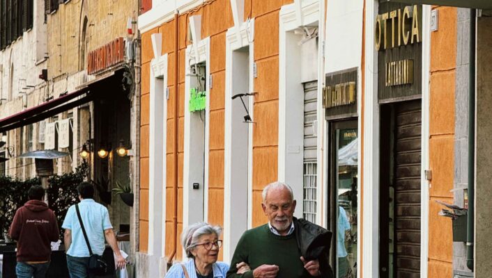 Ottica - An older couple walks arm in arm down a city sidewalk past an Ottica (optics/eyewear) shop, with colorful buildings and other pedestrians in the background.
