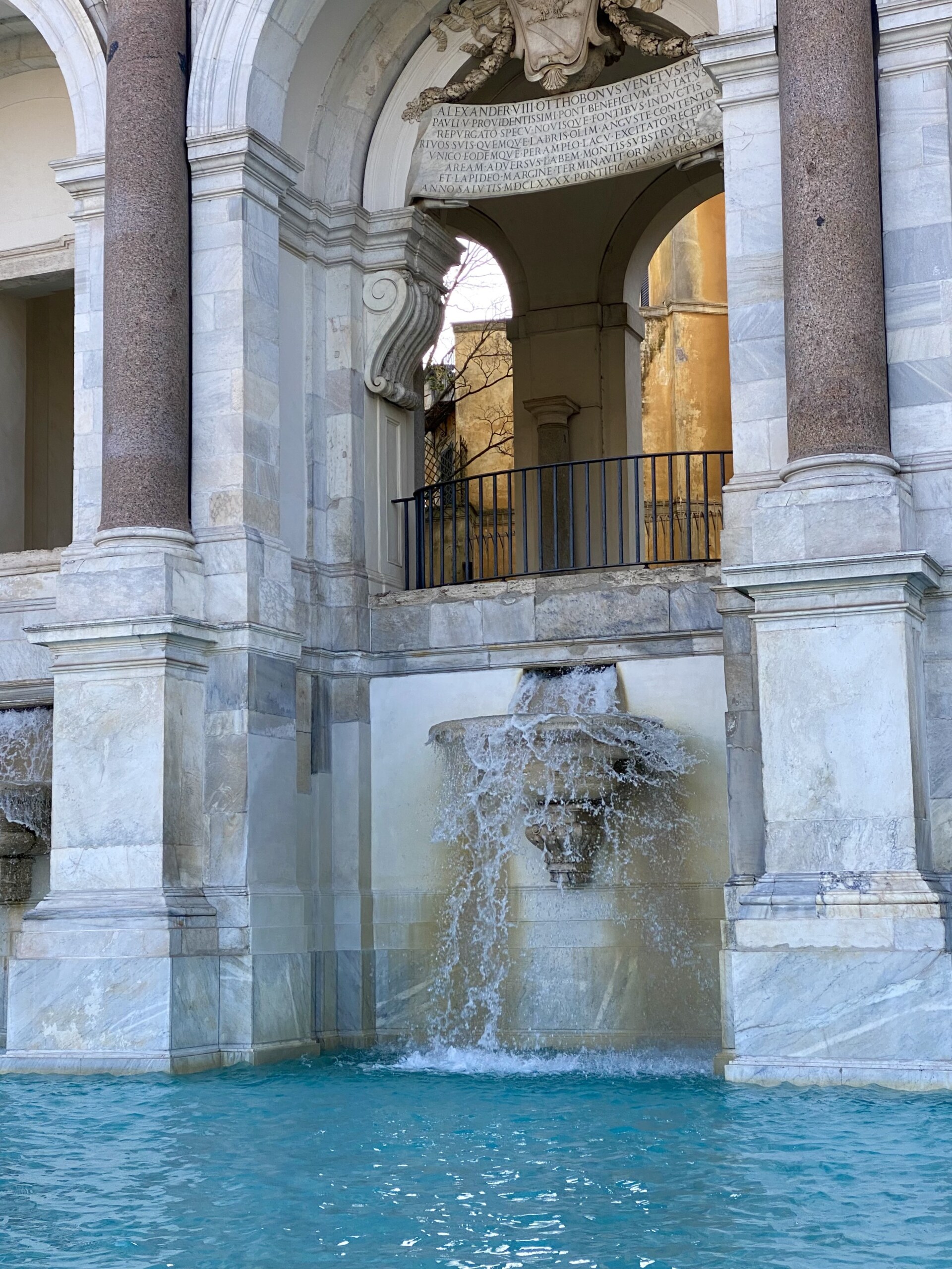 Fontana dell'Acqua Paola A marble fountain with water pouring from a sculpted lions head into a blue pool, set within an ornate stone pavilion featuring arches, columns, and a carved inscription above.