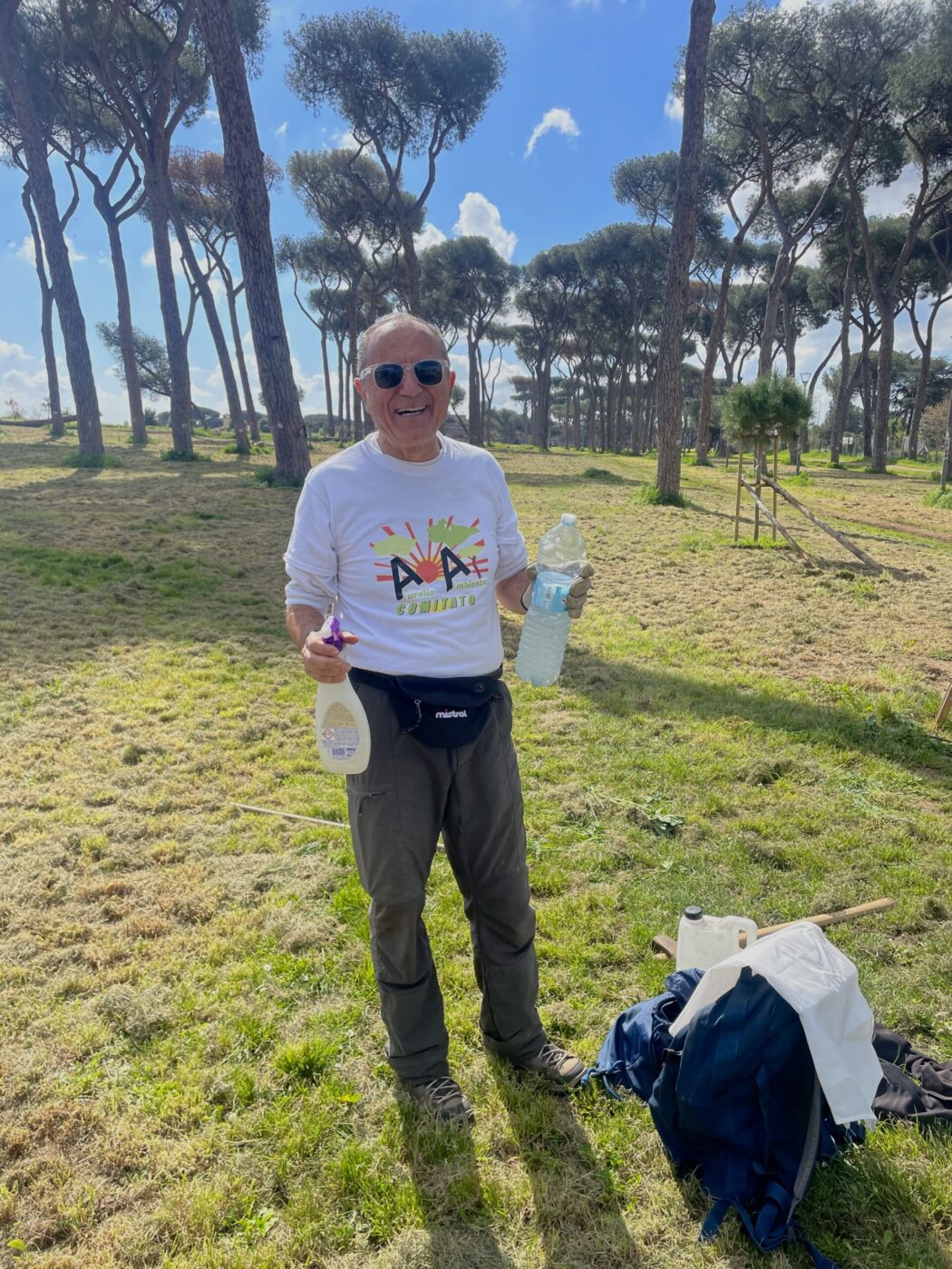 An older man stands smiling in a sunny park, holding a spray bottle and a water bottle. He wears sunglasses, a white t-shirt with colorful graphics, gray pants, and a black waist pouch. Tall trees and scattered clouds are in the background.