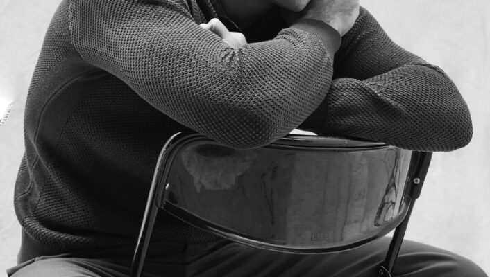 Giuseppe Maggio - A man sits on a backward-facing chair, resting his arms and chin on the chair’s back. He appears thoughtful, gazing slightly upward. The image is in black and white.