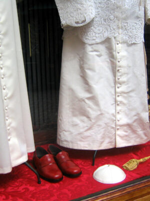 Abiti clericali bianchi - Gli abiti clericali bianchi sono esposti in vetrina con scarpe rosse, uno zucchetto bianco e un cordone dorato su una superficie di velluto rosso. Gli abiti presentano bottoni decorativi e dettagli in pizzo.