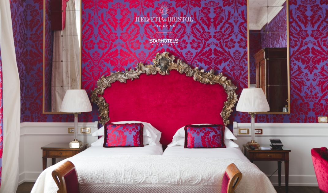 Una lussuosa camera d'albergo: una vivace carta da parati damascata color magenta e rosso fa da sfondo a una testiera decorata in oro e rosso, completata da candide lenzuola, due lampade su comodini in legno e cuscini decorativi coordinati.