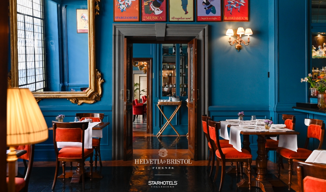Elegante interno del ristorante - Elegante ristorante con pareti blu scuro, sedie arancioni, tovaglie bianche, quadri, un grande specchio con cornice dorata e un lampadario; la vista si estende fino alla zona bar attraverso doppie porte aperte.