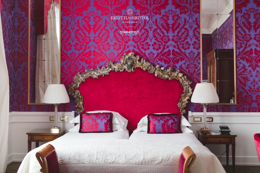 Una lussuosa camera d'albergo: un grande letto con una testiera rossa decorata e bordata in oro è in contrasto con la vivace carta da parati damascata rossa e viola; due comodini con lampade fiancheggiano il letto e le pareti a specchio riflettono l'elegante arredamento della stanza.