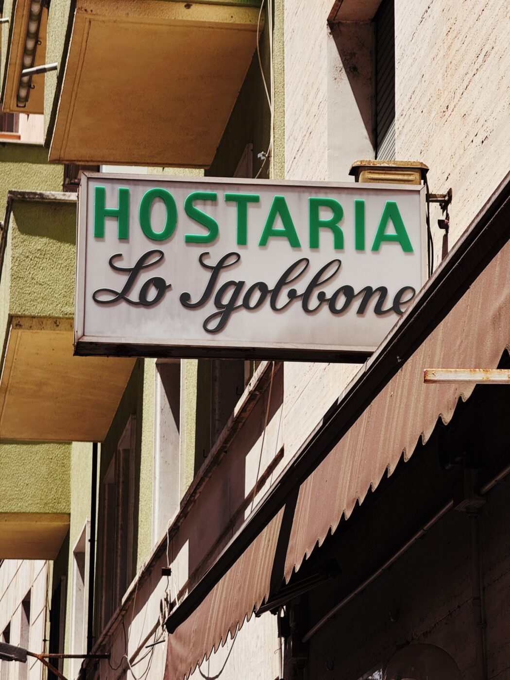HOSTARIA Lo Sgobbone - Un cartello d'epoca con la scritta HOSTARIA Lo Sgobbone è appeso a un edificio, con scritte verdi e nere su sfondo bianco, sopra un marciapiede fiancheggiato da tende da sole.