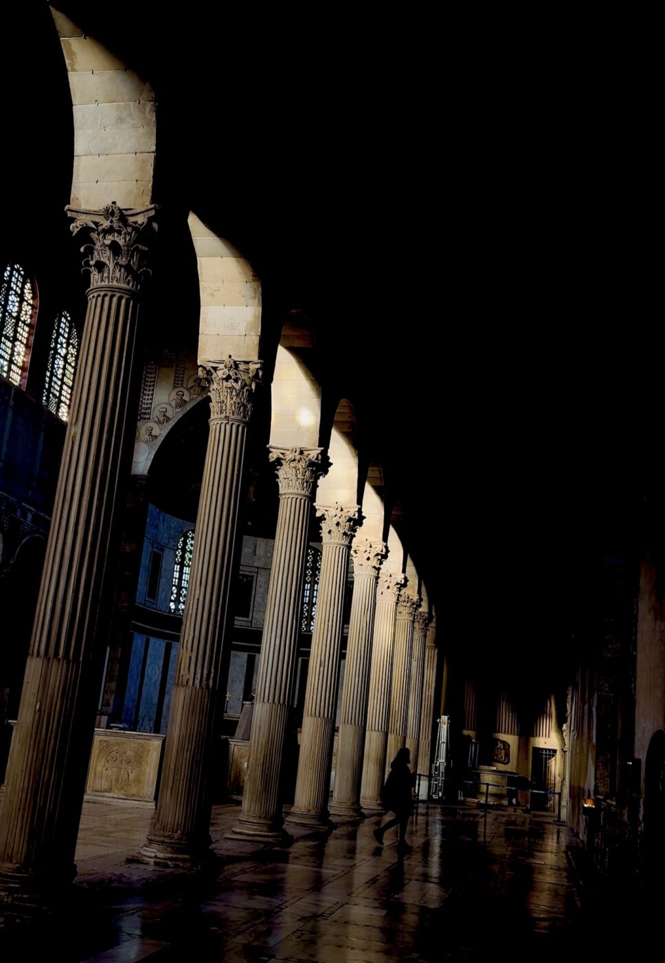 Alte colonne in pietra in una sala storica - Alte colonne in pietra costeggiano l'interno di un edificio storico scarsamente illuminato, la luce del sole filtra attraverso le alte finestre e proietta ombre; una figura solitaria cammina sullo sfondo.