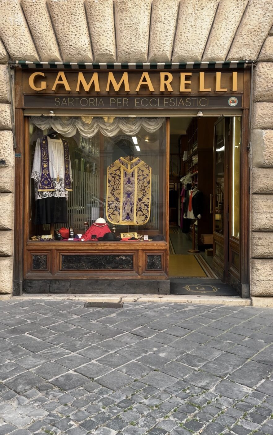 Vetrina di Gammarelli - Vetrina di Gammarelli, una sartoria per abiti ecclesiastici, con paramenti liturgici elaborati in viola e oro esposti nella vetrina, con una strada acciottolata di fronte.