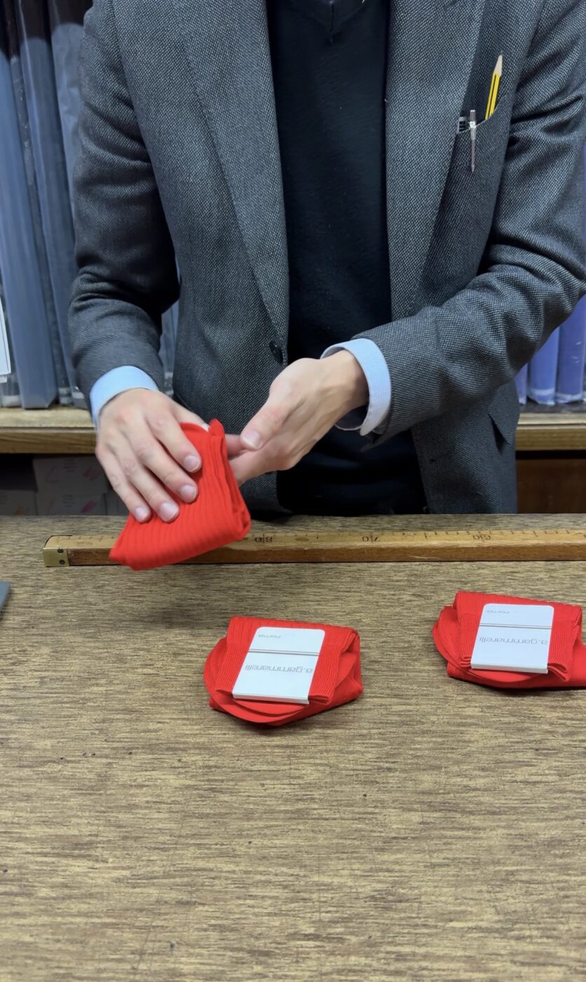 [Esposizione di calzini rossi BRAMAMARTE] - Una persona con una giacca grigia piega un paio di calzini rosso acceso su un tavolo di legno, con altri due paia di calzini rossi con l'etichetta BRAMAMARTE disposti ordinatamente lì vicino.