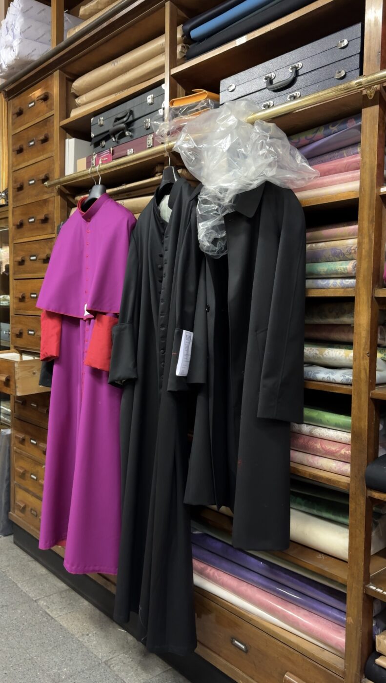 Tre abiti ecclesiastici (una tonaca viola con polsini rossi, una tonaca nera e un cappotto nero) sono appesi davanti a scaffali di legno pieni di rotoli di tessuto e cassetti in una sartoria.