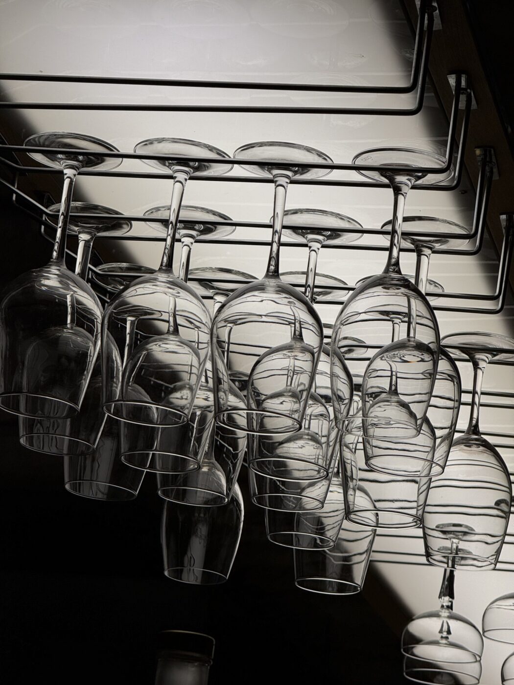 A row of empty wine glasses hangs upside down from a metal rack, illuminated by overhead lighting that casts reflections and shadows on the glasses.