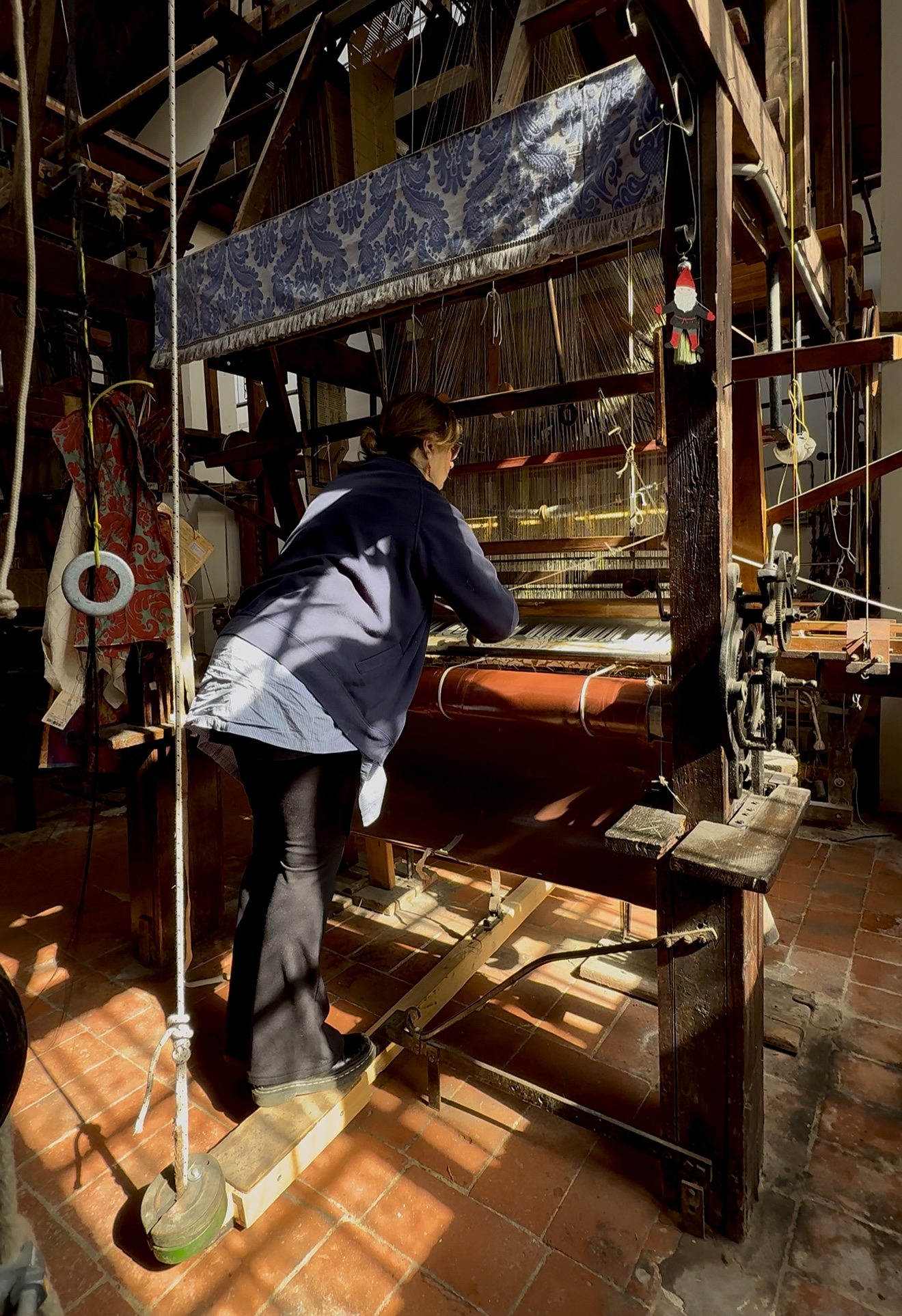 Una persona è in piedi davanti a un grande telaio di legno, intenta a tessere in una stanza illuminata dal sole, con pavimenti piastrellati e vari strumenti appesi nelle vicinanze. La luce del sole filtra da una finestra, proiettando ombre sulla scena.