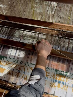 A persons hand operates a traditional wooden loom, weaving a fabric with a floral pattern in green and yellow. Sunlight casts shadows across the threads and the persons arm.
