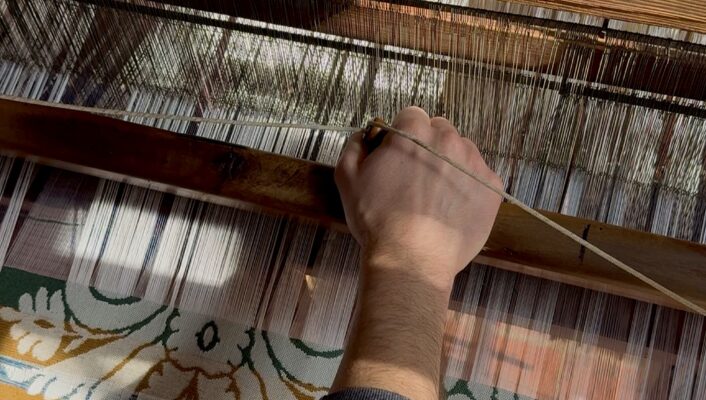 A persons hand operates a traditional wooden loom, weaving a fabric with a floral pattern in green and yellow. Sunlight casts shadows across the threads and the persons arm.