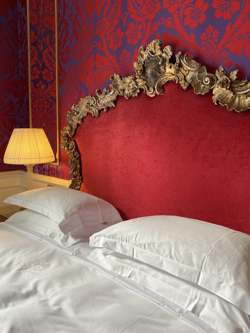 Un letto lussuoso con lenzuola bianche e una testiera in velluto rosso con elaborati dettagli dorati, accanto a una lampada da tavolo accesa, contro una parete con carta da parati floreale rossa e viola.