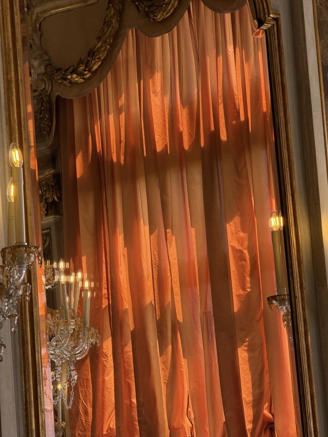 [Nessun titolo immagine] - Un grande specchio decorato con cornice dorata riflette i lampadari scintillanti e le tende drappeggiate color pesca, delicatamente illuminati da una luce calda in un elegante interno classico.