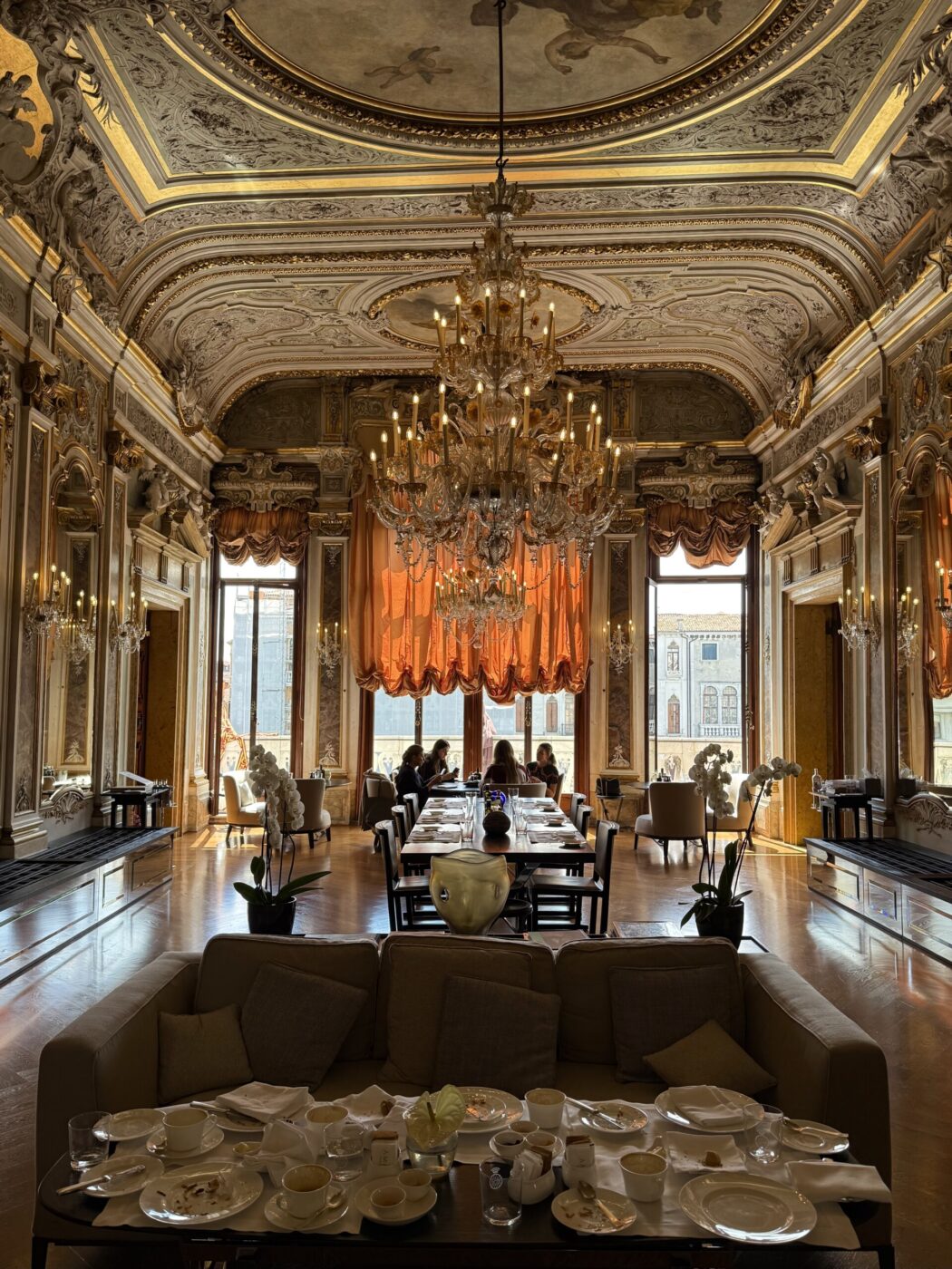 [Nessun titolo dell'immagine] - Elegante sala con soffitti decorati in oro, lampadari, alte finestre drappeggiate con tende arancioni e lunghi tavoli da pranzo apparecchiati per il tè. In primo piano, divani e un tavolo sono coperti da tazze e piatti da tè.