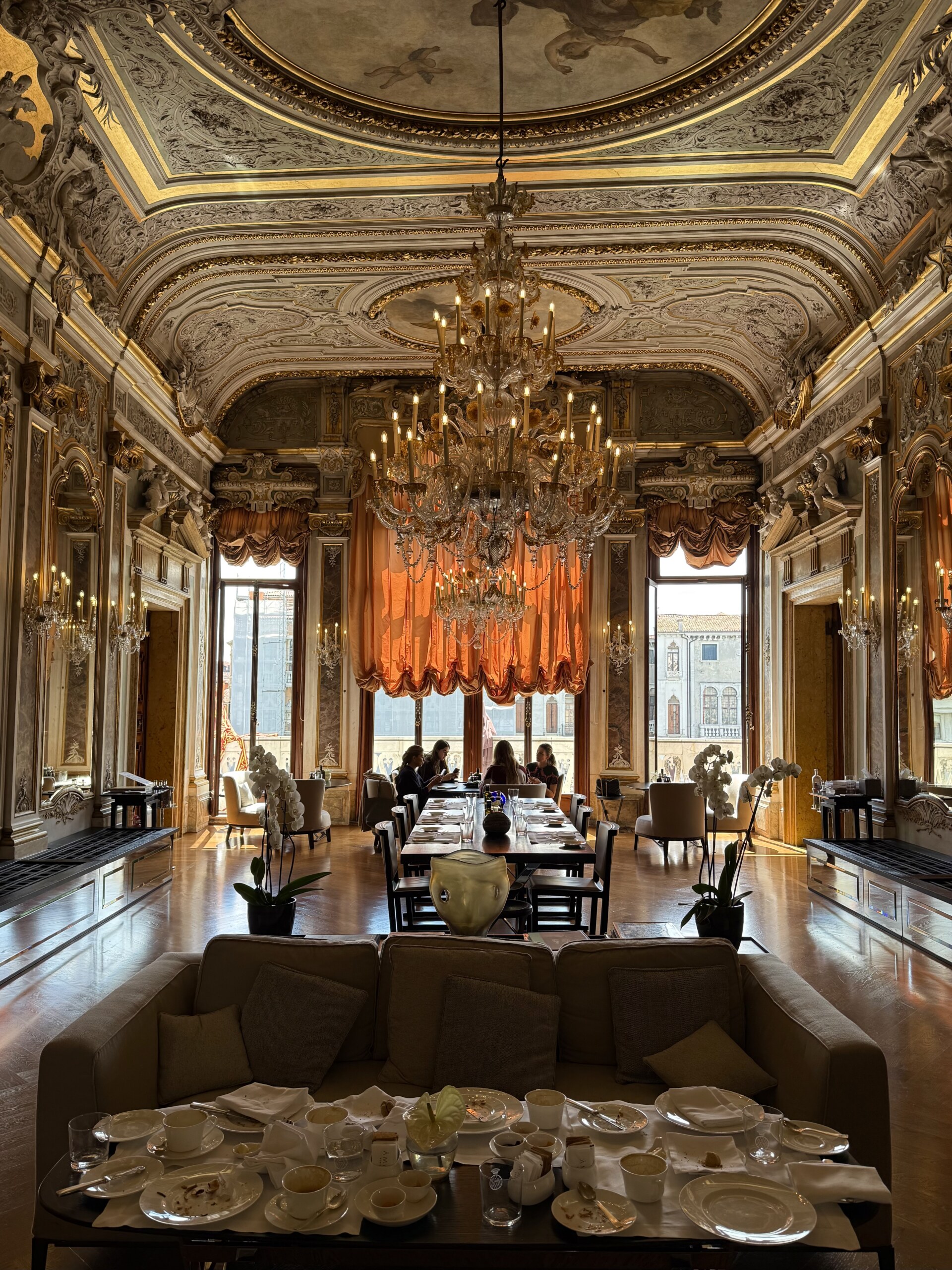 [Nessun titolo dell'immagine] - Elegante sala con soffitti decorati in oro, lampadari, alte finestre drappeggiate con tende arancioni e lunghi tavoli da pranzo apparecchiati per il tè. In primo piano, divani e un tavolo sono coperti da tazze e piatti da tè.