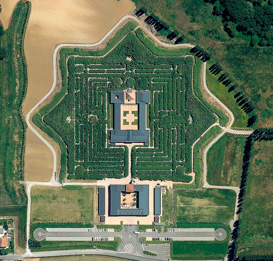 Vista aerea di un labirinto di giardini a forma di stella con siepi che circondano un edificio centrale, immerso tra campi e alberi, con un vialetto d'accesso e un parcheggio in basso.