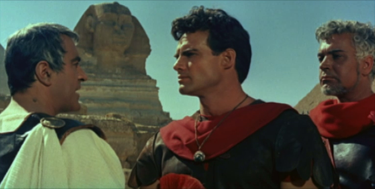 Three men in ancient Roman attire stand in front of the Sphinx in Egypt, having a serious conversation under a clear blue sky. The central man wears a red cloak, and pyramids are visible in the background.