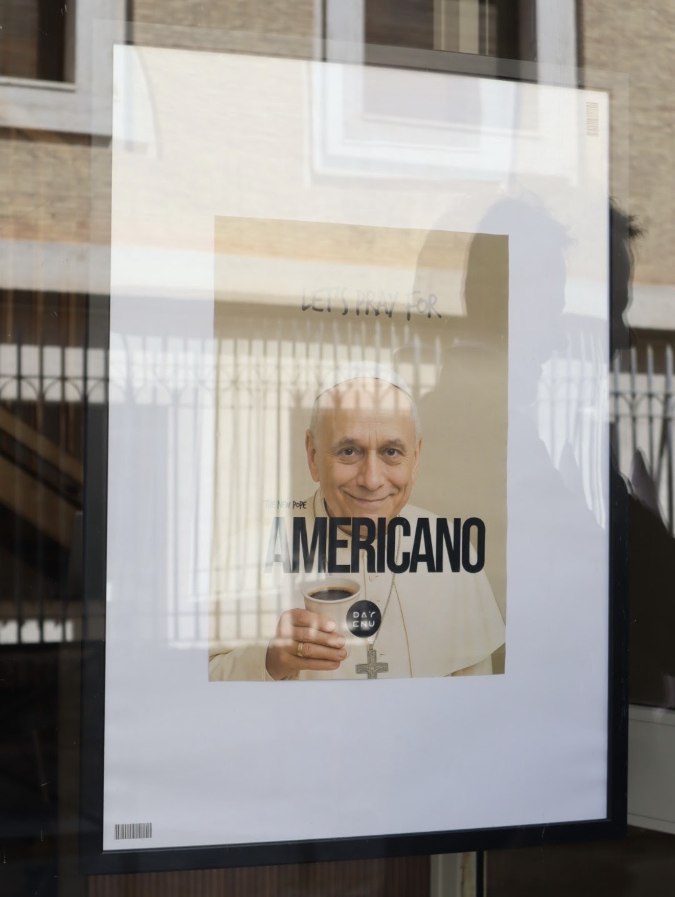 LET’S PRAY FOR AMERICANO - A framed poster shows a man in papal attire holding a cup of coffee, with the words “LET’S PRAY FOR AMERICANO” printed across the image. The photo is taken through a reflective glass window.