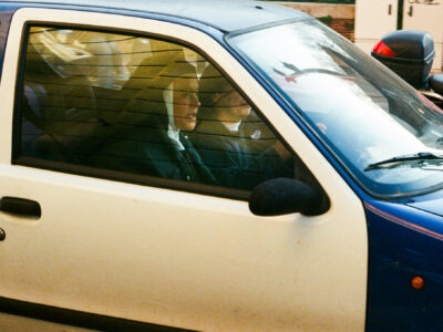 Two nuns in car - Two nuns dressed in habits sit in the front seats of a small blue and white car, with the driver focused on the road. Sunlight creates reflections on the window, partially obscuring their faces.