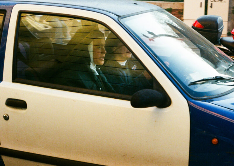Two nuns in car - Two nuns dressed in habits sit in the front seats of a small blue and white car, with the driver focused on the road. Sunlight creates reflections on the window, partially obscuring their faces.