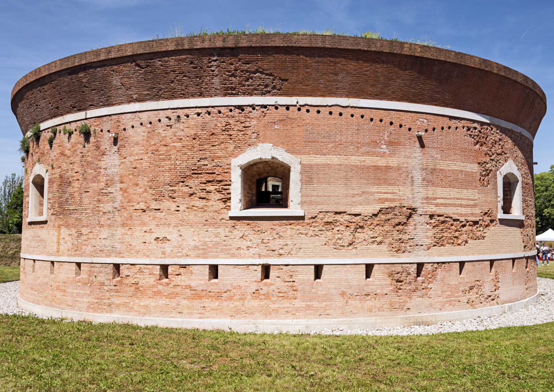 [Titolo mancante] - Una grande fortezza rotonda di mattoni con piccole finestre e mura segnate dal tempo sorge su un prato sotto un cielo azzurro. Si vedono delle persone in lontananza vicino a una tenda gialla.