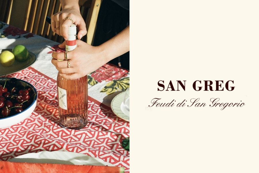 SAN GREG Feudi di San Gregorio - A person’s hands open a bottle of rosé wine on a table with a red-patterned cloth, surrounded by cherries, watermelon, apples, and plates. The right side reads: “SAN GREG Feudi di San Gregorio.”.