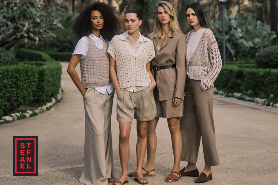 Collezione Primavera Stefanel - Quattro donne sono all'aperto in un giardino, indossando abiti primaverili Stefanel dai toni neutri, tra cui top in maglia, camicette, pantaloni ampi, un abito blazer e sandali. Il logo Stefanel appare nell'angolo in basso a sinistra.
