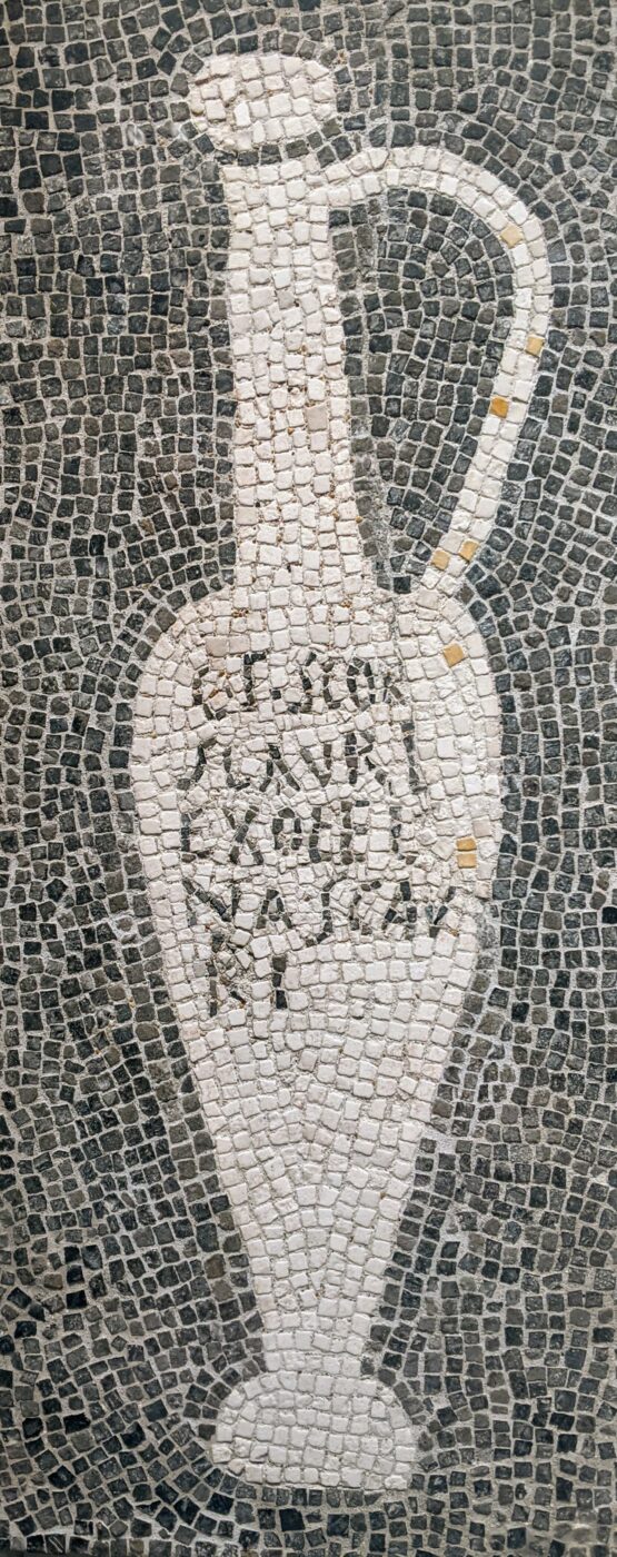 [Titolo dell'immagine] - Un mosaico raffigura un'anfora bianca con un manico su sfondo scuro, con testo latino disposto verticalmente all'interno. Le tessere del mosaico sono piccole e di forma irregolare.
