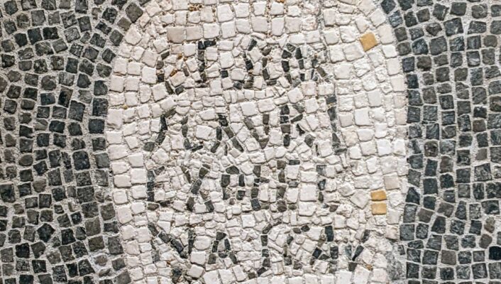 [Titolo dell'immagine] - Un mosaico raffigura un'anfora bianca con un manico su sfondo scuro, con testo latino disposto verticalmente all'interno. Le tessere del mosaico sono piccole e di forma irregolare.
