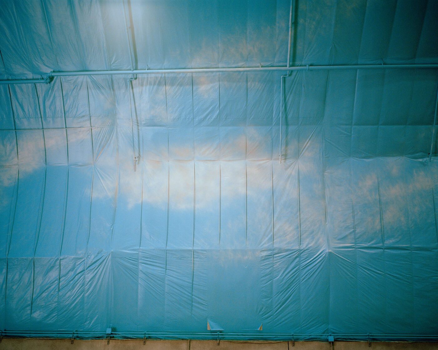 Blue Plastic Sky_Rome_Blue plastic sheeting covers a ceiling, with light and shadows creating the illusion of clouds drifting across a sky. Pipes and seams run along the ceiling, adding texture and structure to the scene.
