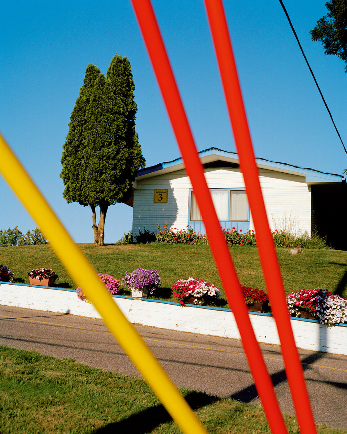 A small white house_Rome_A small white house with the number 3 on it sits on a grassy hill, with colorful flowers in front and a tall green tree beside it. Bright red and yellow poles are visible in the foreground. The sky is clear and blue.