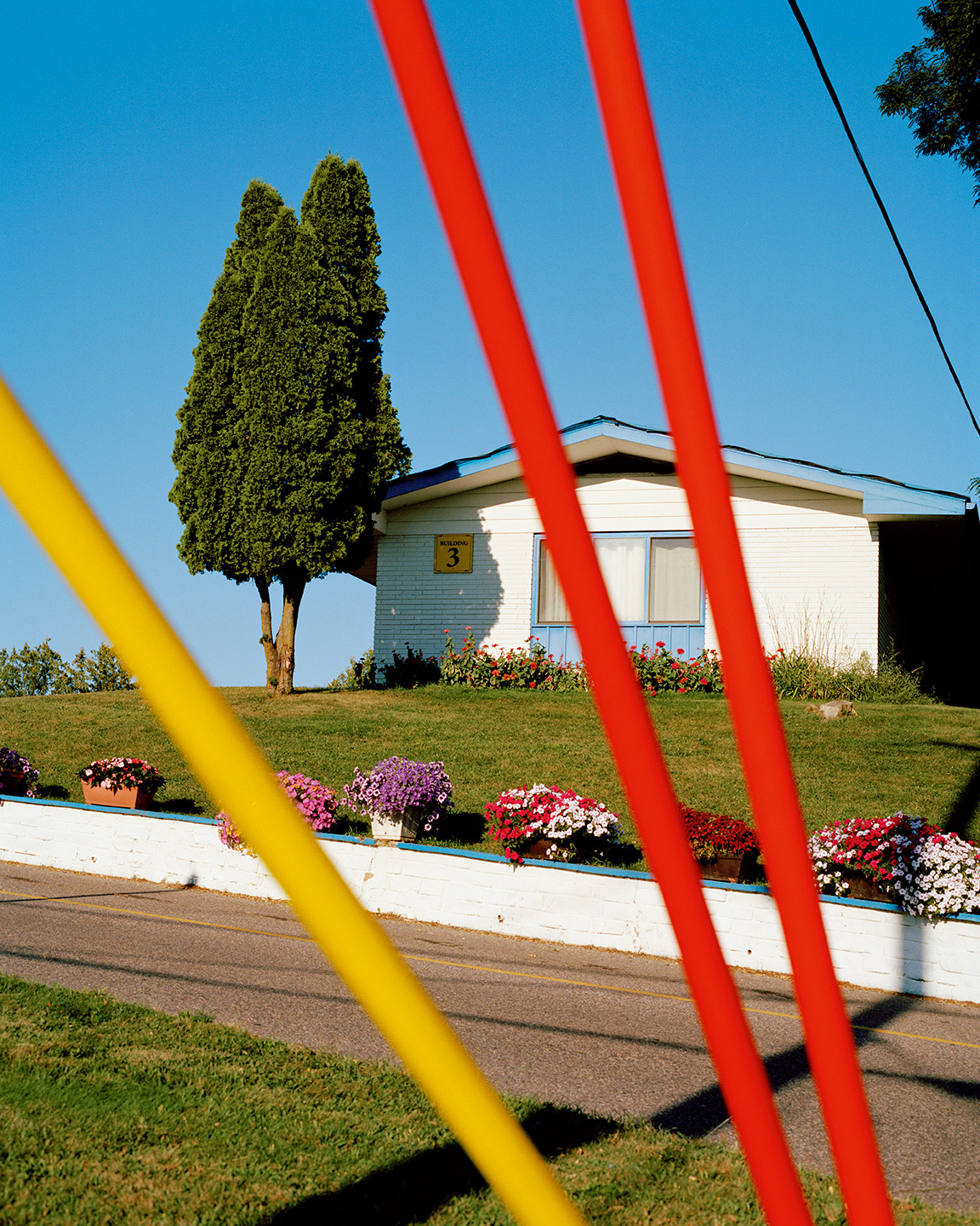 A small white house_Rome_A small white house with the number 3 on it sits on a grassy hill, with colorful flowers in front and a tall green tree beside it. Bright red and yellow poles are visible in the foreground. The sky is clear and blue.