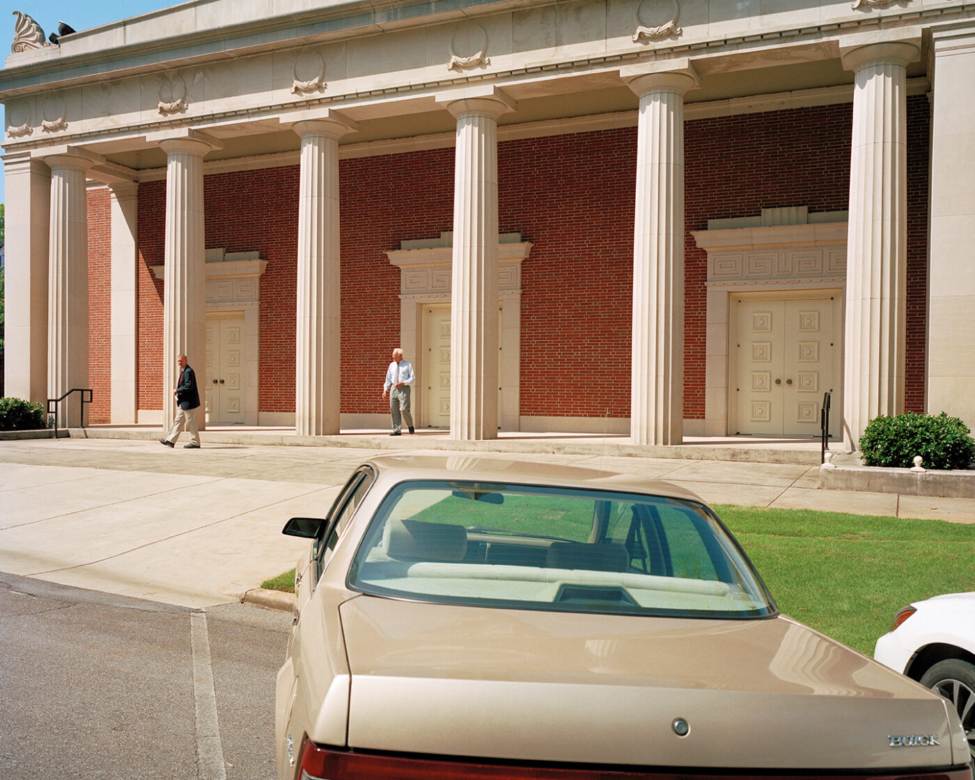 Car Parked_Rome_A tan car is parked in front of a red brick building with four tall white columns. Two people are walking on the sidewalk near the building’s entrance doors. Bright sunlight casts shadows on the scene.