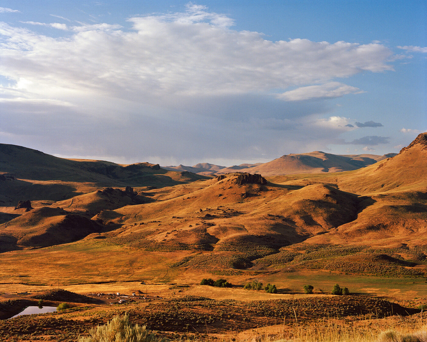 Rolling Golden Hills Landscape_Rome_Rolling golden hills and rugged rock formations under a partly cloudy sky, with sunlight casting shadows across the landscape and a small pond and trees in the foreground.