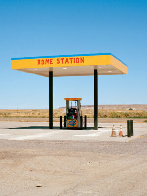 STAZIONE DI SERVIZIO DI ROMA - Una singola pompa di benzina si erge sotto una pensilina gialla e blu con la scritta "STAZIONE DI SERVIZIO DI ROMA" in un paesaggio deserto e aperto, sotto un cielo azzurro e terso. Due coni stradali sono posizionati vicino alla pompa e sullo sfondo si vedono colline in lontananza.