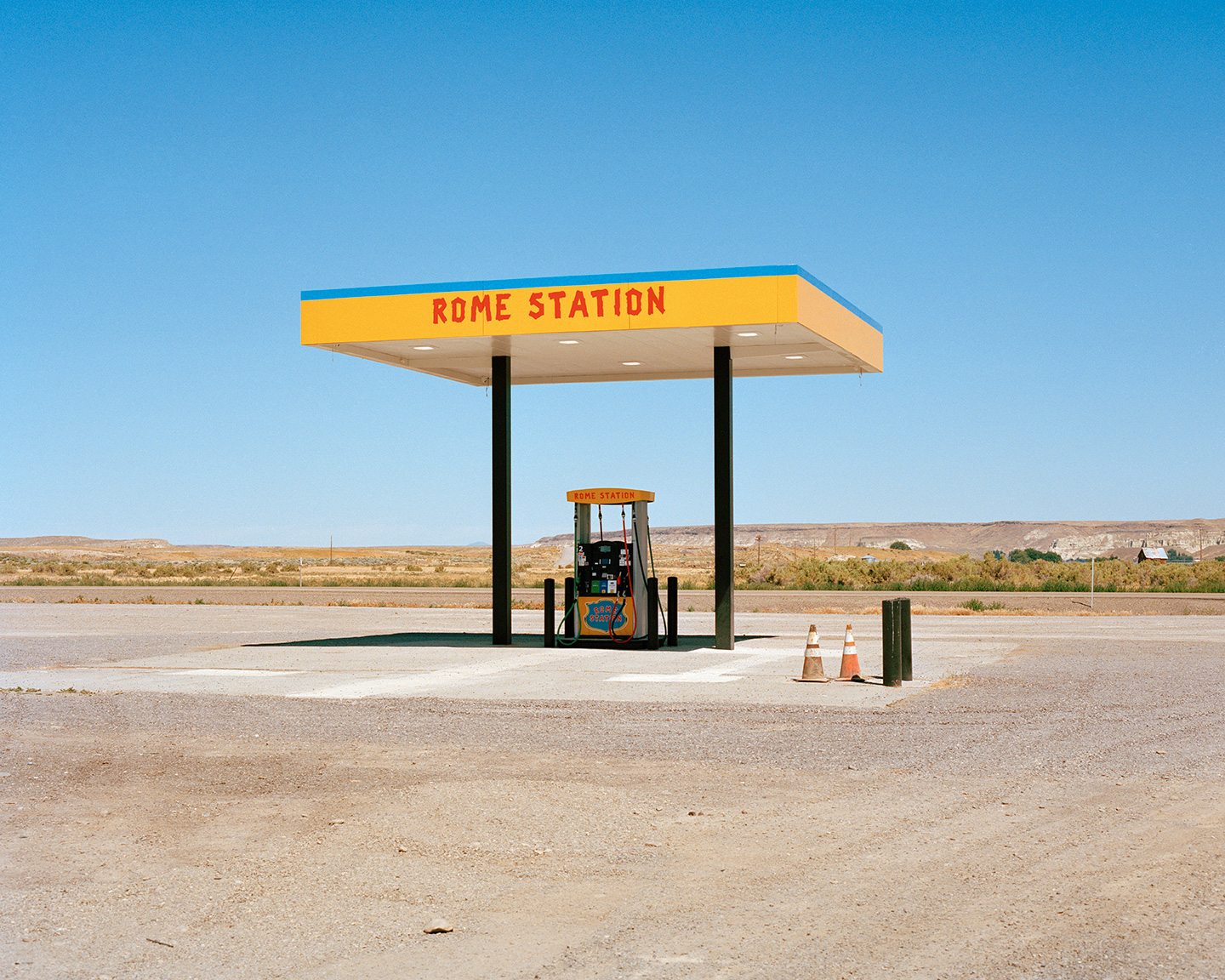 Rome Station_A single yellow and blue gas pump stands under a canopy labeled “ROME STATION” in red letters, surrounded by an empty, arid landscape under a clear blue sky. Two traffic cones are nearby.