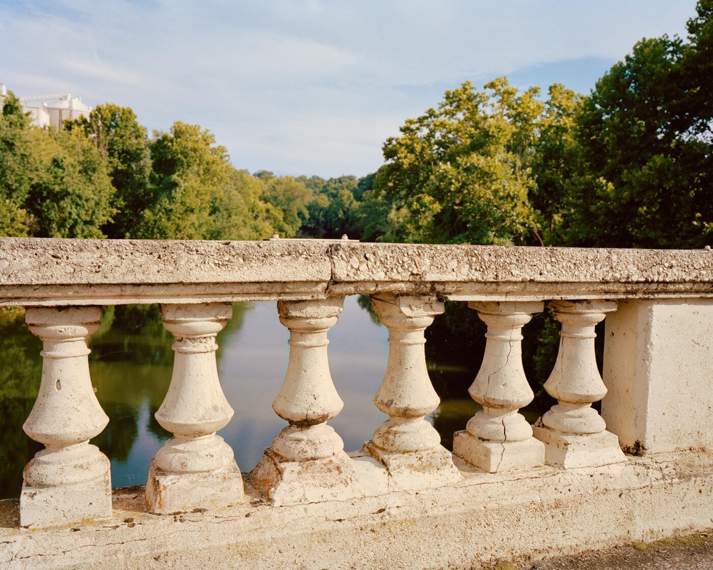 Riverside Serenity_Rome_A stone balustrade overlooks a calm river surrounded by lush green trees under a blue sky, with sunlight casting warm tones on the scene.