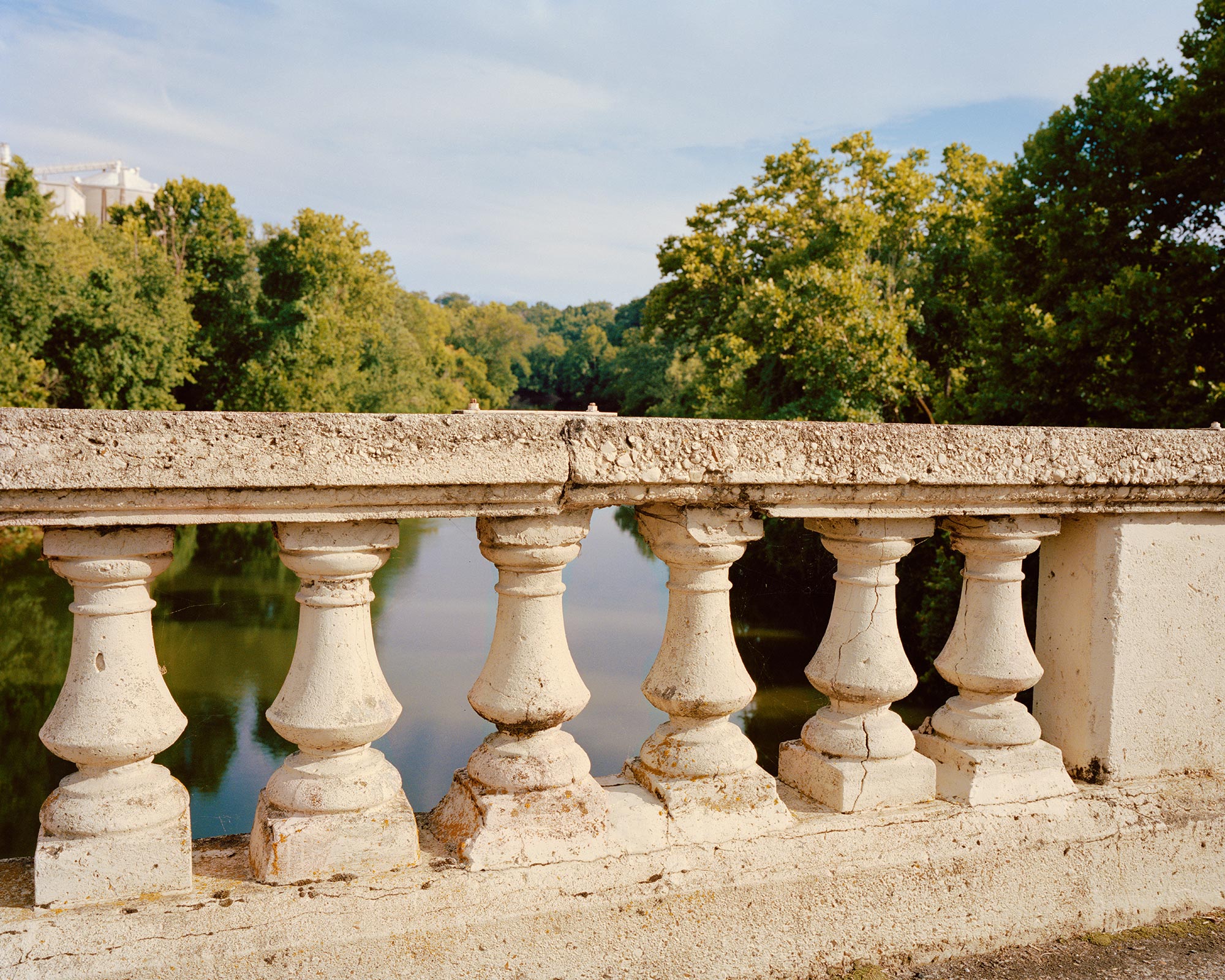 Riverside Serenity_Rome_A stone balustrade overlooks a calm river surrounded by lush green trees under a blue sky, with sunlight casting warm tones on the scene.