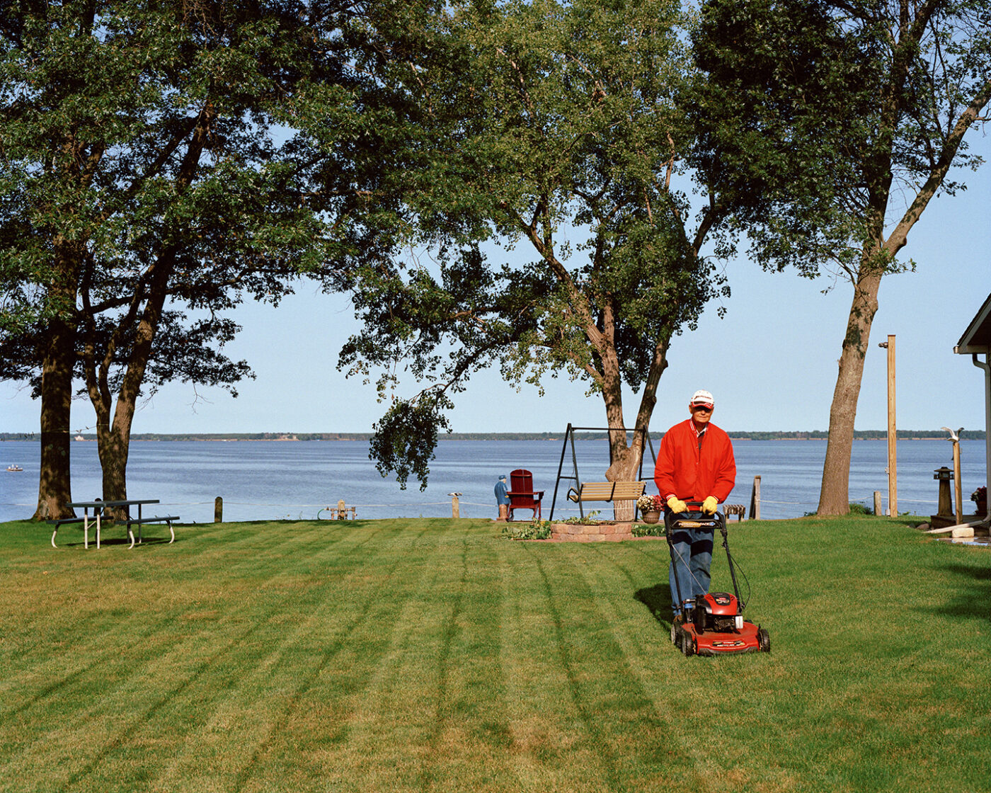 Lakeside Lawncare_Rome_A person in a red jacket and hat mows a grassy lawn near a lake, with trees, a swing, and Adirondack chairs in the background under a clear blue sky.